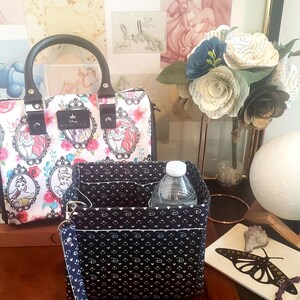 May include: A white handbag with a floral and princess design, featuring a black handle and a matching organizer. The organizer is black with a white pattern and contains a water bottle. A bouquet of paper flowers and a decorative tray are also visible.