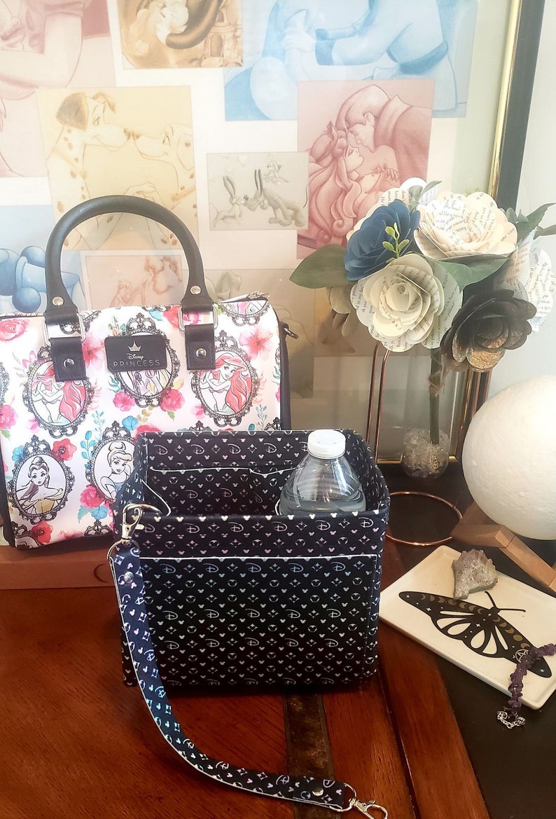 May include: A white handbag with a floral and princess design, featuring a black handle and a matching organizer. The organizer is black with a white pattern and contains a water bottle. A bouquet of paper flowers and a decorative tray are also visible.
