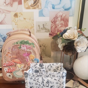 May include: A light pink Alice in Wonderland themed backpack with a colorful illustration of Alice, the Cheshire Cat, and mushrooms. A white and black patterned organizer sits in front of the backpack. Artwork of Disney characters is in the background.