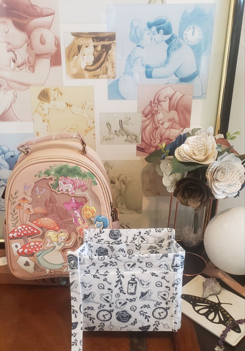 May include: A light pink Alice in Wonderland themed backpack with a colorful illustration of Alice, the Cheshire Cat, and mushrooms. A white and black patterned organizer sits in front of the backpack. Artwork of Disney characters is in the background.