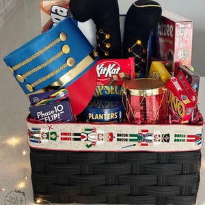 May include: A black woven basket filled with a blue and red toy soldier hat, black plush boots, a red toy drum, and various board games including Jenga, Yahtzee, Phase 10 Flip, and Uno, plus a KitKat bar and Planters peanuts, wrapped in nutcracker ribbon.