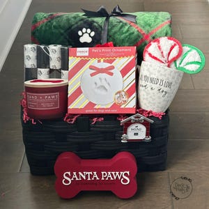 Könnte beinhalten: Ein schwarzer geflochtener Korb mit einer grünen karierten Decke, einer roten Sand + Paws Kerze, einem Reece Pet's Print Ornament Set, einem Becher mit der Aufschrift All You Need Is Love And A Dog, einem Hundehütten-Anhänger und einem roten Knochenschild.