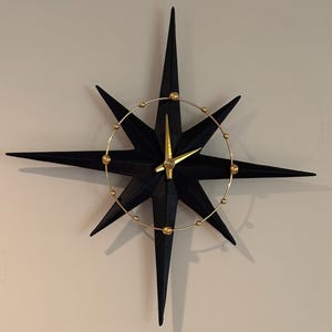 May include: A black starburst clock with gold-colored accents. The clock features a star-shaped design with pointed arms and a circular gold ring with small gold spheres. The clock hands are gold-colored.
