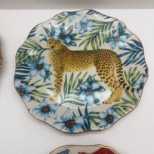 Puede incluir: Un plato decorativo con un dise&ntilde;o de guepardo. El plato tiene un fondo blanco con patrones florales azules y hojas verdes. El guepardo es amarillo con manchas negras. El plato tiene un borde dorado.