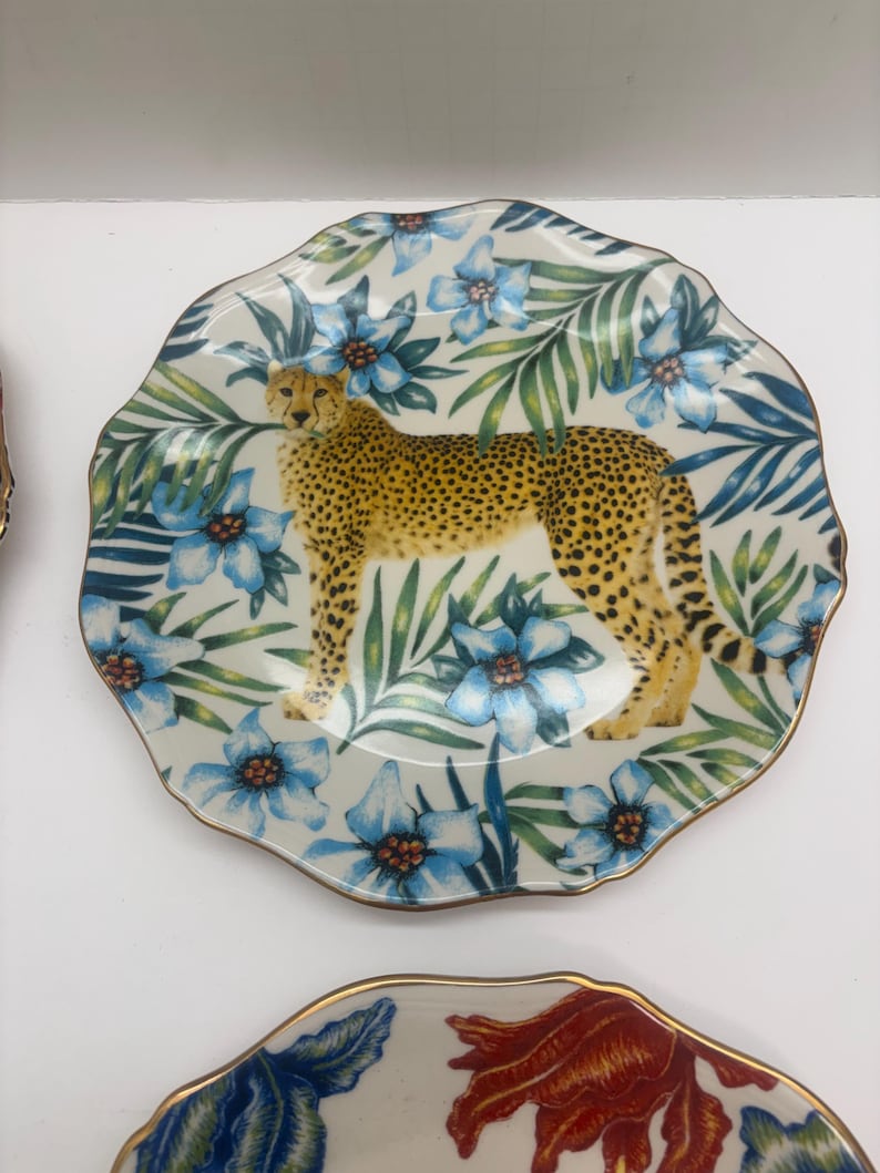 Puede incluir: Un plato decorativo con un dise&ntilde;o de guepardo. El plato tiene un fondo blanco con patrones florales azules y hojas verdes. El guepardo es amarillo con manchas negras. El plato tiene un borde dorado.