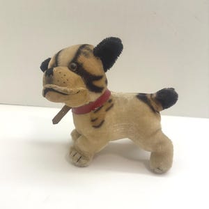 May include: A vintage plush dog toy in tan and black. The dog has black ears, a red collar, and black stripes on its face and body. The toy is standing on a white surface.