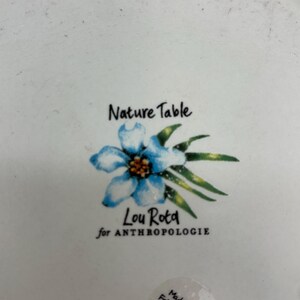 Puede incluir: Plato de cer&aacute;mica blanco con dise&ntilde;o floral y el texto "Nature Table" y "Lou Rota for ANTHROPOLOGIE". El plato presenta una ilustraci&oacute;n de flor azul con hojas verdes.