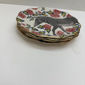Puede incluir: Una pila de platos decorativos con forma ovalada. Cada plato presenta una ilustraci&oacute;n de cebra y un dise&ntilde;o floral con flores rojas, hojas verdes y un borde dorado. Los platos est&aacute;n apilados.