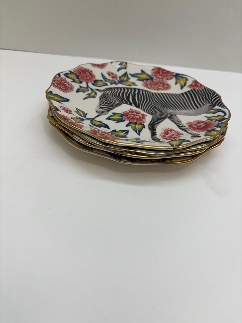 Puede incluir: Una pila de platos decorativos con forma ovalada. Cada plato presenta una ilustraci&oacute;n de cebra y un dise&ntilde;o floral con flores rojas, hojas verdes y un borde dorado. Los platos est&aacute;n apilados.