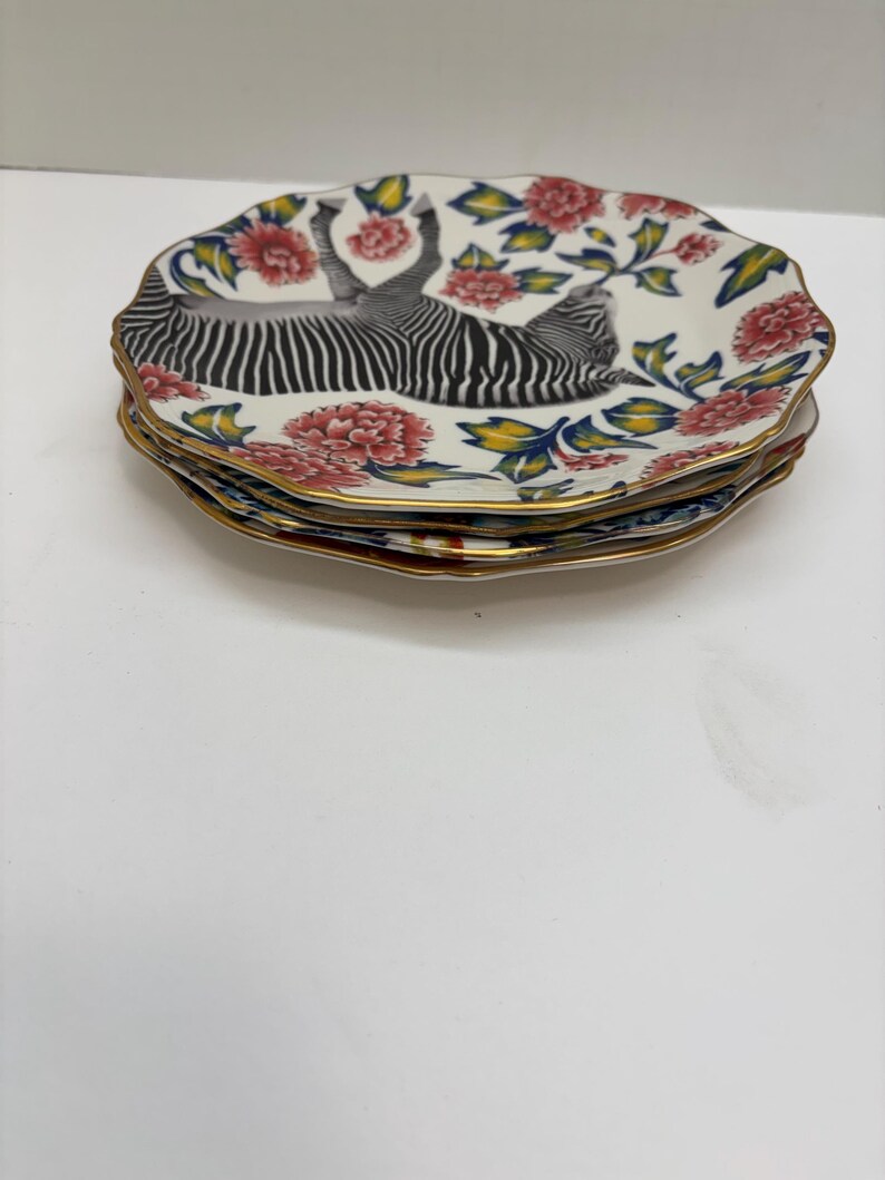 Puede incluir: Una pila de cuatro platos decorativos. Cada plato presenta un dise&ntilde;o de cebra en blanco y negro, rodeado de flores rojas, hojas verdes y un borde dorado. Los platos tienen un borde festoneado.