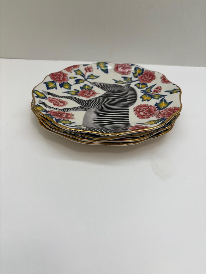 Puede incluir: Una pila de platos decorativos con un dise&ntilde;o floral y de cebra. Los platos tienen un fondo blanco con flores rojas y rosas, hojas verdes y un borde dorado. El centro de cada plato presenta una ilustraci&oacute;n de cebra.