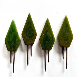 May include: Four vintage green diamond-shaped hat pins with dark brown metal pins. The hat pins have a marbled appearance with shades of green and brown. The pins are arranged in a row against a white background.