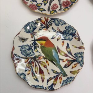 Puede incluir: Platos decorativos de cer&aacute;mica con borde festoneado. Uno presenta un p&aacute;jaro colorido posado en una rama con detalles florales azules. Otro muestra un dise&ntilde;o de cebra con detalles florales rojos. Ambos platos tienen un borde dorado.