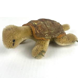 May include: A small turtle figurine crafted from tan fuzzy pipe cleaners with a textured brown shell featuring gold-colored accents. The turtle has four legs with pointed tips and a rounded head with a small dark eye visible on the side.