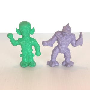 May include: Two small, plastic monster figures. One is green with large ears and a mask-like face. The other is purple, with a reptilian appearance and muscular arms. Both are standing and appear to be toys or collectibles.