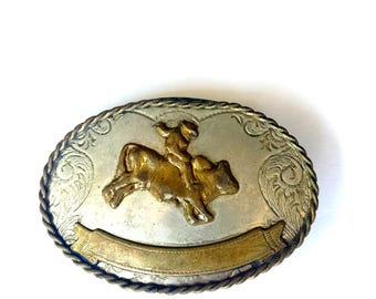 May include: Oval-shaped silver and gold belt buckle featuring a rodeo scene. The buckle has a gold-colored rider on a bull in the center, with a silver background and a decorative border. A gold banner is at the bottom.