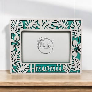 May include: White wooden picture frame with a teal green background and a cutout design of tropical leaves and flowers. The frame says "Hawaii" at the bottom and "Koko Blue Designs" in the centre.