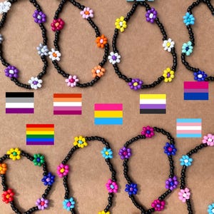 Peut inclure: Bracelets de perles avec des fleurs colorées sur un cordon noir. Les bracelets présentent des fleurs roses, jaunes et bleues. Des drapeaux de la fierté, dont les drapeaux arc-en-ciel, transgenre et bisexuel, sont également affichés.