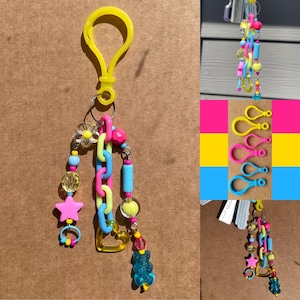 Op de afbeelding: Een kleurrijke sleutelhanger met een gele clip, met een ketting van roze en blauwe schakels, een roze ster, een doorzichtige bloem en een blauwe gummybeer-bedel. De sleutelhanger heeft diverse kralen en een geel hart.