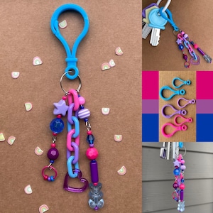 Op de afbeelding: Een blauwe sleutelhangerclip met een kralenbedel. De bedel bevat een paarse ster, blauwe en roze kralen, een hart en een gummybeer. Extra sleutelhangers en sleutelhangers met sleutels zijn ook afgebeeld.