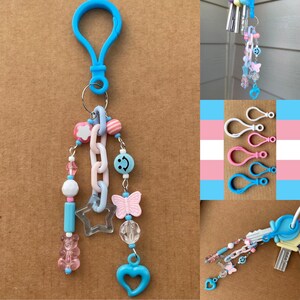 Op de afbeelding: Een blauwe sleutelhanger met een karabijnhaak, versierd met een ketting, kralen en bedels. De bedels zijn onder meer een gummybeer, een ster, een vlinder, een smiley en een hart. Het kleurenpalet is pastelroze, blauw en wit.