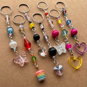 May include: Seven colorful beaded keychains with different charms. The charms include a butterfly, a heart, a crystal, and a rainbow-colored cylinder. The keychains are made with silver-toned metal rings and chains.