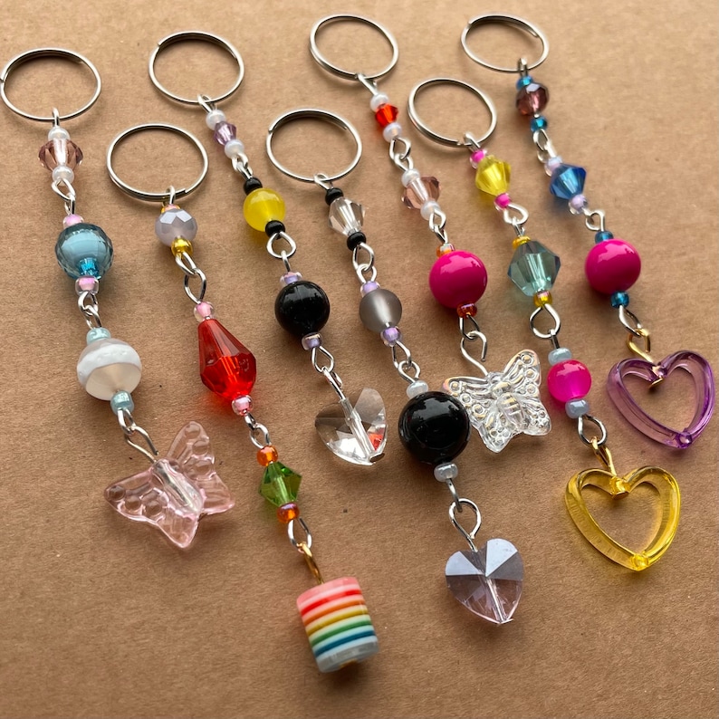 May include: Seven colorful beaded keychains with different charms. The charms include a butterfly, a heart, a crystal, and a rainbow-colored cylinder. The keychains are made with silver-toned metal rings and chains.
