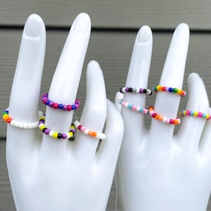 May include: A set of colorful beaded rings, including a white ring, a rainbow ring, a purple, pink, and black ring, and a ring with alternating black and white beads.