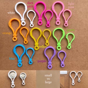 Op de afbeelding: Een verzameling kleurrijke karabijnhaak sleutelhangers in verschillende maten. De sleutelhangers zijn wit, oranje, geel, blauw, paars, donkerroze, lichtroze en groen. De afbeelding bevat ook de tekst "small vs large".