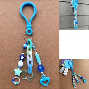 Op de afbeelding: Een blauwe sleutelhanger met een karabijnhaak, een sleutelring en decoratieve bedels. De bedels omvatten blauwe en witte kralen, ster- en hartvormen en een schakelkettingontwerp. De sleutelhanger is een mix van blauw en helder plastic.