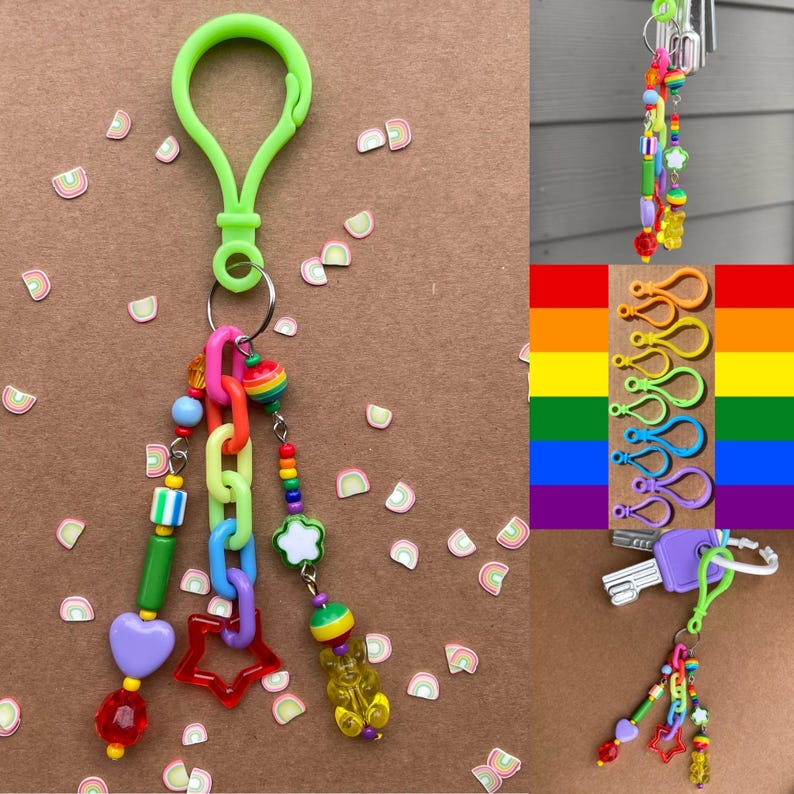Op de afbeelding: Een kleurrijke sleutelhanger met een groene clip, met een verscheidenheid aan kralen, waaronder een hart, een ster en een gummybeer. De sleutelhanger heeft regenboogkleurige accenten en een kleine regenboogvormige bedel. De achtergrond is lichtbruin.