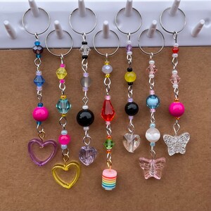 May include: Seven colorful beaded keychains with different charms. The charms include hearts, butterflies, and a rainbow-colored cake. The keychains are made with various beads, including clear, pink, blue, yellow, and green.