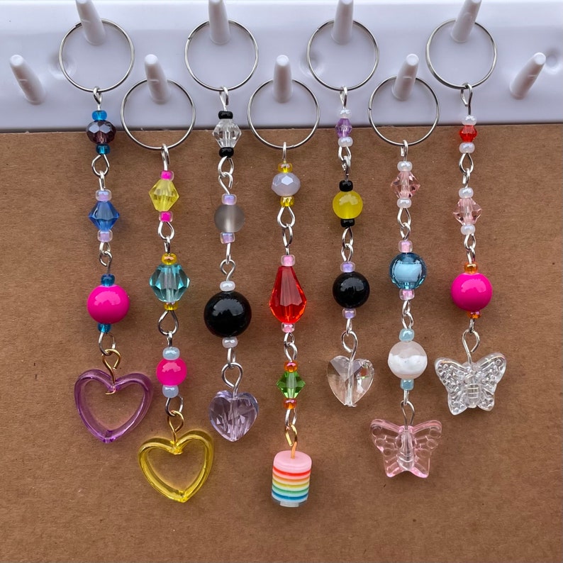 May include: Seven colorful beaded keychains with different charms. The charms include hearts, butterflies, and a rainbow-colored cake. The keychains are made with various beads, including clear, pink, blue, yellow, and green.