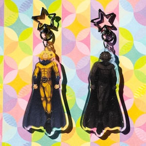 May include: Two acrylic keychains featuring superhero characters. One keychain depicts a character in a yellow and black costume with a blue cape. The other keychain shows a character in a black costume with a black cape. Both keychains have a star-shaped clasp.