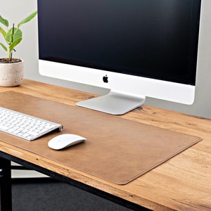 Op de afbeelding: Een bruine leren bureau mat met een wit toetsenbord en muis op een houten bureau met een computermonitor en een plant in een pot.