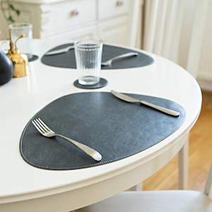 May include: A white table set with two gray oval placemats, each with a silver fork and knife. A glass of water sits on one of the placemats.