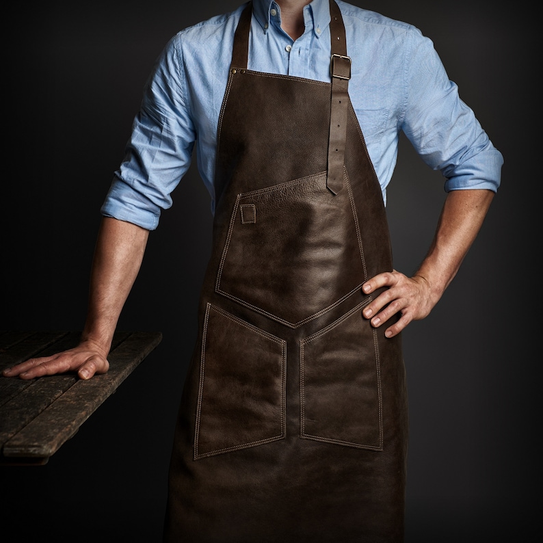 May include: A brown leather apron with multiple pockets and adjustable straps. The apron is worn by a person with a light blue button-down shirt.