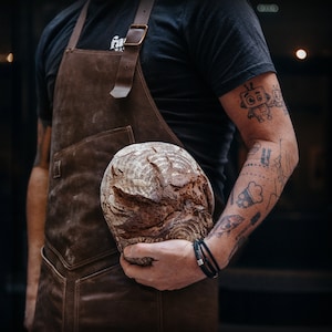 May include: A person wearing a brown leather apron with a large, round loaf of bread in their hand. The apron has a strap that goes over the shoulder and a buckle. The person has tattoos on their arm.