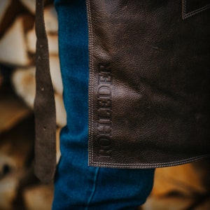May include: A brown leather apron with the word "ROHFEDER" embossed in white stitching. The apron is worn over blue jeans.