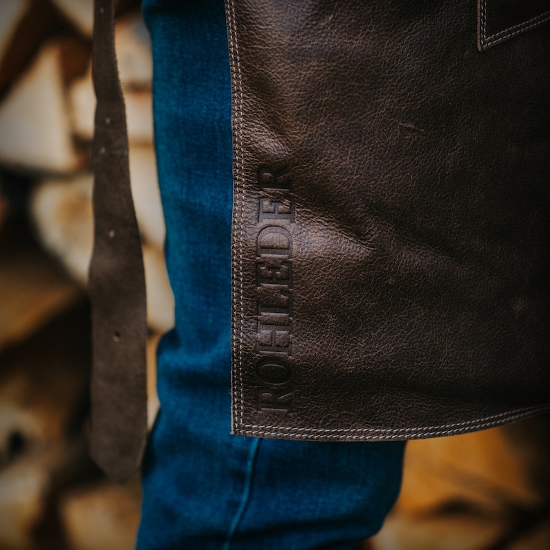 May include: A brown leather apron with the word "ROHFEDER" embossed in white stitching. The apron is worn over blue jeans.