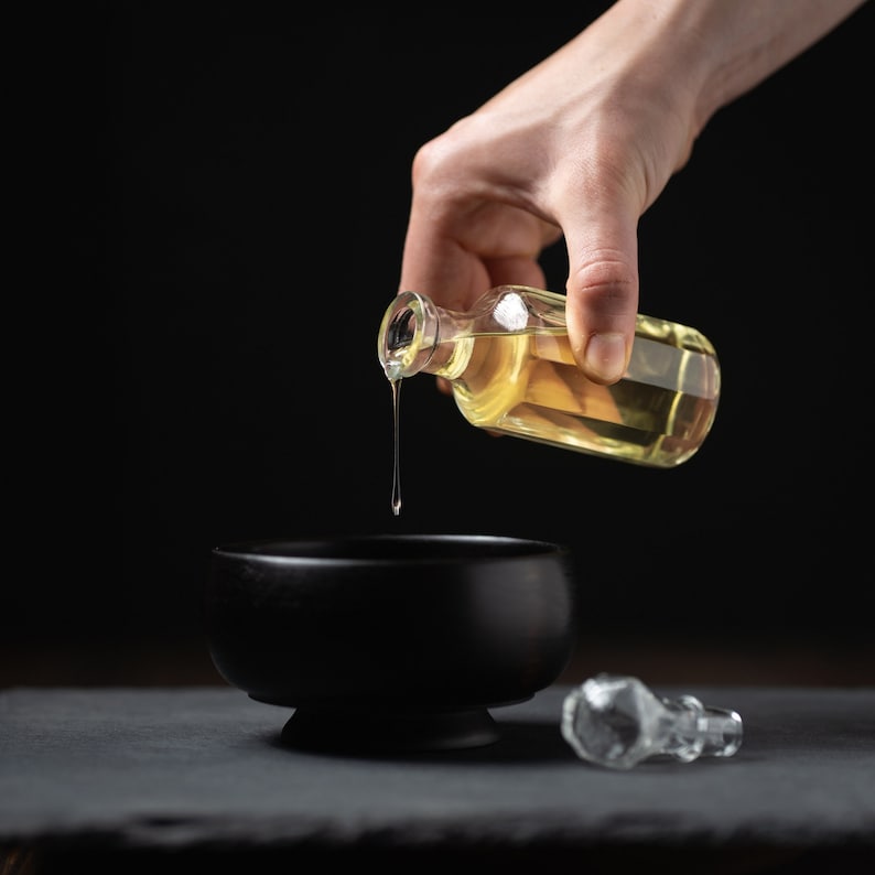 May include: A hand pouring golden liquid from a clear glass bottle into a black bowl. The bowl is sitting on a dark gray surface.