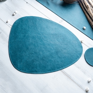 Könnte beinhalten: Tischset aus Leder in Teal-Farbe, oval geformt, auf einer weißen Holzoberfläche.