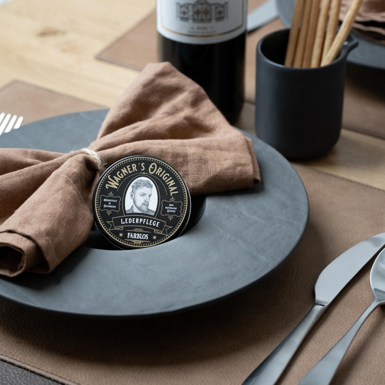 May include: A black and gold round tin with the text "Wagner's Original" and "Lederpflege Farblos" on it. The tin is sitting on a black plate with a brown linen napkin.