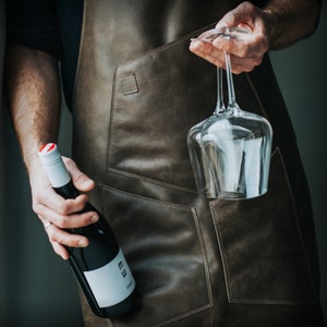 May include: A person wearing a brown leather apron holds a bottle of wine and a clear wine glass. The bottle has a white label with the text "EW" and "Zweigelt".