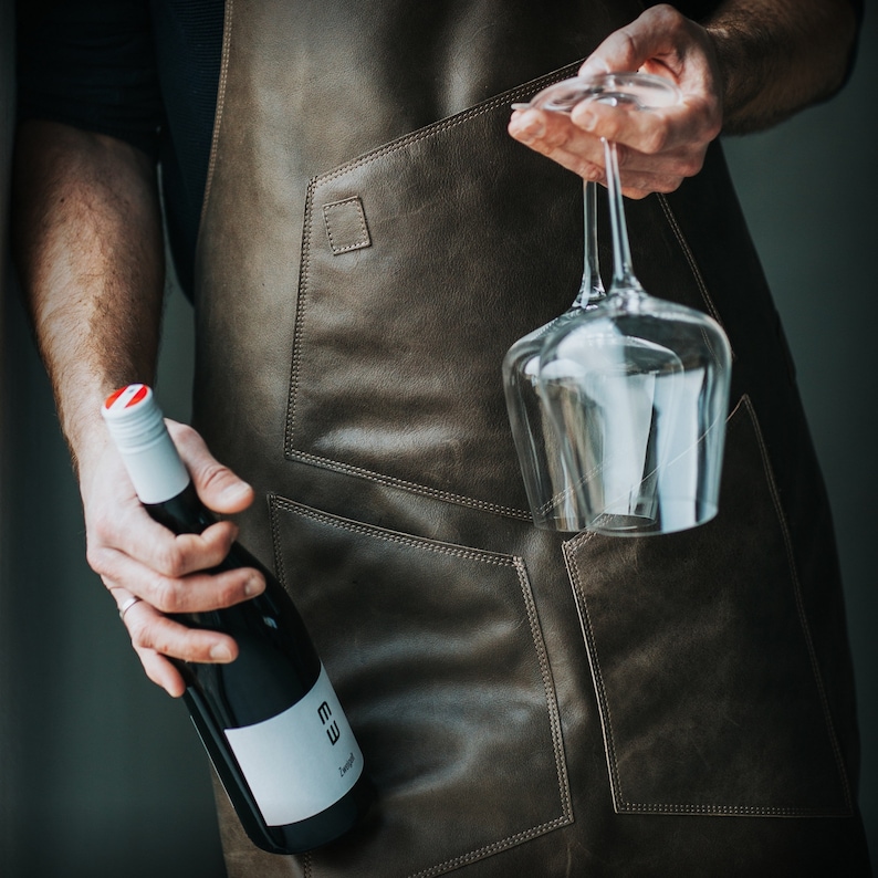 May include: A person wearing a brown leather apron holds a bottle of wine and a clear wine glass. The bottle has a white label with the text "EW" and "Zweigelt".