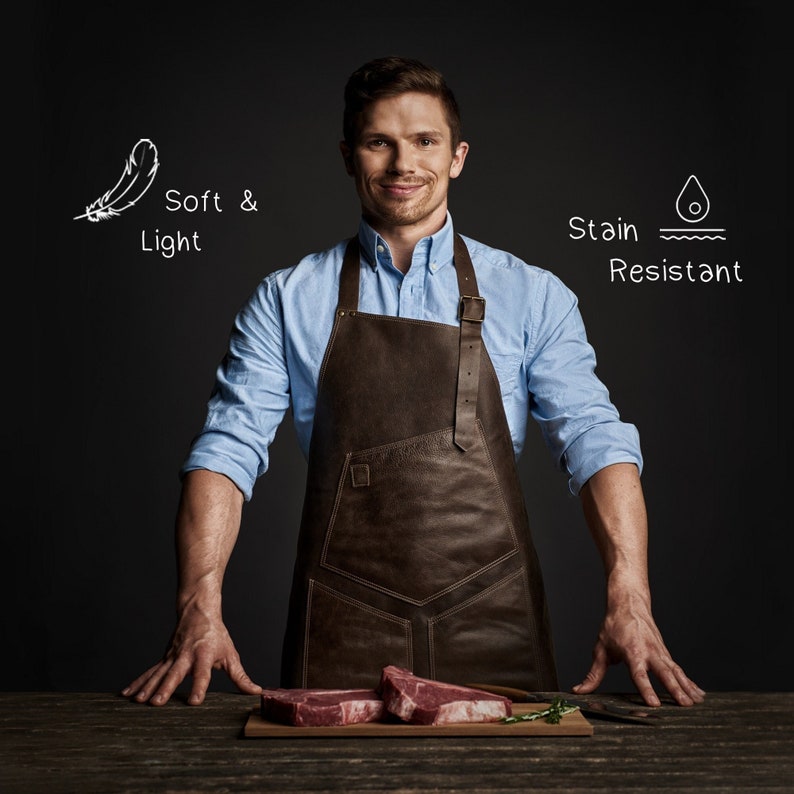 May include: A man wearing a brown leather apron stands in front of a dark background. He is smiling and looking at the camera. There are two raw steaks on a wooden cutting board in front of him. The text "Soft & Light" and "Stain Resistant" is written on the background.