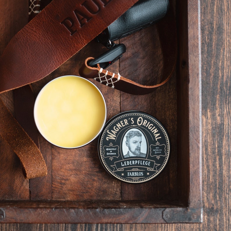 May include: A small tin of leather care product with a black and gold label. The label says "Wagner's Original" and "Lederpflege Farblos". The tin is sitting in a wooden box with a brown leather strap.