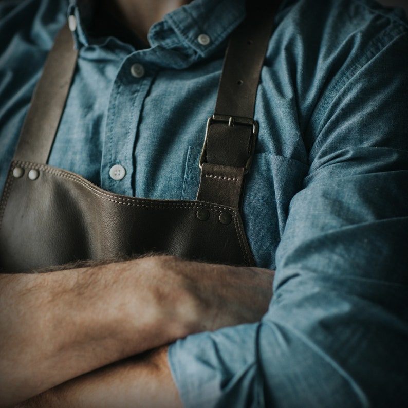 May include: A person wearing a blue denim shirt with a brown leather apron. The apron has a metal buckle and is fastened with straps.
