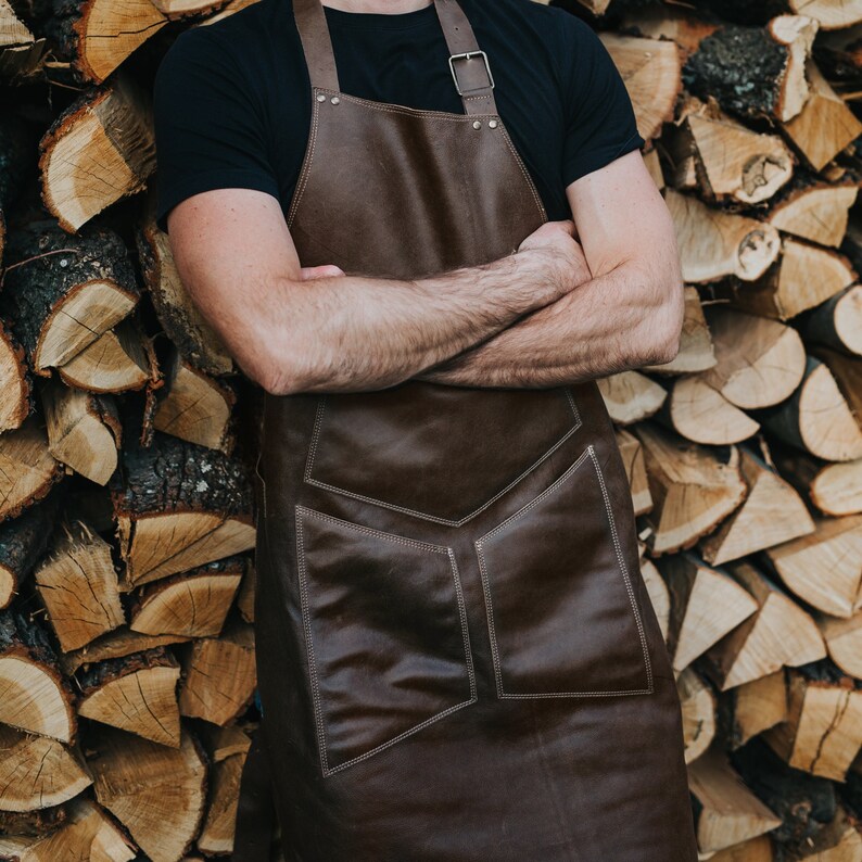 May include: A brown leather apron with adjustable straps and a large pocket. The apron is worn by a person with their arms crossed.