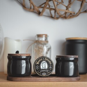 May include: A black and gold round container with the text "Wagner's Original" and "Lederpflege Farblos" on it. The container is sitting on a wooden surface with two black ceramic jars with wooden lids.